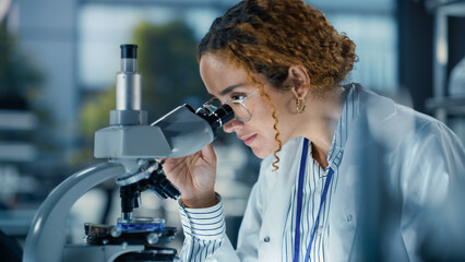 Wall Mural - Hisplanic Female Bioengineer Looking at DNA Samples Under a Microscope in a Modern Applied Science Laboratory. Portrait of a Young Lab Engineer in White Coat Inventing New Medical Drugs