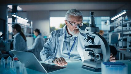 Leinwandbilder - Senior Medical Research Scientist Looking at Biological Samples Under a Microscope in an Applied Science Laboratory. Portrait of a Handsome Middle Aged Doctor Working on a Research Project on a Laptop