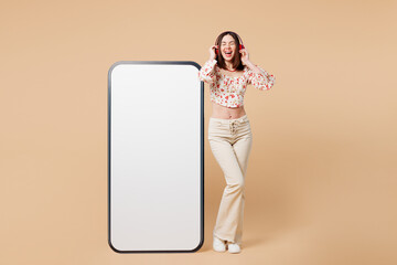 Poster - Full body young woman wear white top casual clothes big huge blank screen mobile cell phone smartphone with area listen to music in headphones isolated on plain beige background. Lifestyle concept.