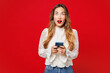 © ViDi Studio - Young shocked surprised happy Caucasian woman she wear white blouse casual clothes hold in hand use mobile cell phone look camera isolated on plain red background studio portrait. Lifestyle concept.