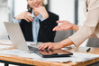 © kenchiro168 - Two businesswomen discussing work at desk with laptop and tablet, showcasing collaboration and teamwork in modern office