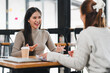 © kenchiro168 - Two women having friendly conversation at table in modern office space, with one holding coffee cup and other gesturing expressively, creating collaborative and engaging moment