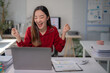 © Tj - Happy young office worker celebrating business success while looking at a laptop, raising hands in excitement at a modern desk filled with financial documents