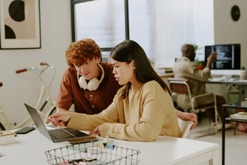 Female and male colleagues sitting at table and looking at monitor while young adult brunette woman showing presentation of brand new project