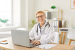 © Studio Romantic - Portrait of a mature senior happy smiling female doctor sitting at the desk working in medical office and looking cheerful at camera. Woman physician wearing stethoscope in clinic using laptop