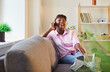 © Studio Romantic - Portrait of happy cheerful african american woman wearing casual clothes sitting on sofa in living room at home talking by smartphone and laughing. Girl resting on couch using mobile phone.