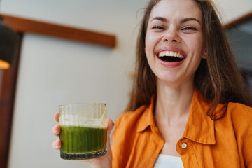 Canvas Print - Smiling young woman with long hair holding a green smoothie, wearing an orange jacket, showcasing healthy eating and vibrant lifestyle in a cozy indoor setting