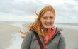 © ullision - Red-haired woman smiles warmly against a breezy beach background, her hair dancing in the wind. The overcast sky and serene coastline evoke a sense of calm and connection to nature