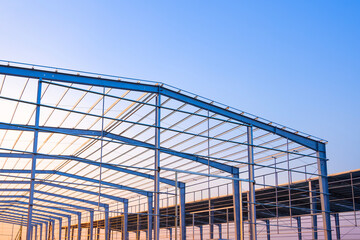 Wall Mural - Factory building framework with metal post and roof beam structure  in construction site against evening sky background, low angle view