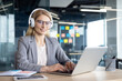© Liubomir - A professional woman wearing headphones works on her laptop at a desk in a modern office. She appears focused and engaged, exemplifying productivity and communication in a business environment.