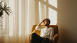 © Bonsales - Young man relaxing in an armchair by the window, soaking up the warm sunlight and savoring a serene moment of calm