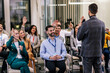 © Mediteraneo - Young business man lecturing his multiethnic colleagues. Having a speech while holding a microphone. Everyone is listening and some are raising their hand.