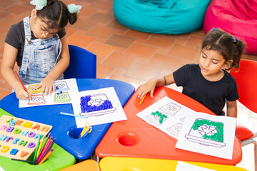 Dos niñas latinas de primaria realizando trabajos sobre el reciclaje en el aula de clases, con mesas coloridas