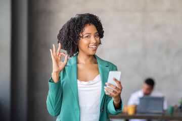 Leinwandbilder - A team member in a corporate office smiles while holding a smartphone and giving an okay gesture. Collaboration thrives in this diverse environment, showcasing successful teamwork.