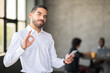 © Prostock-studio - A professional man in a white shirt holds a smartphone while making an 'OK' gesture. He appears confident and engaged in a collaborative office environment with colleagues working in the background.