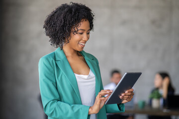 Leinwandbilder - A woman smiles while interacting with a tablet in a contemporary office setting. Background features a diverse team engaged in discussions, showcasing teamwork and innovation in business.