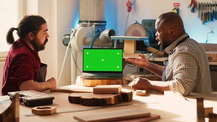 BIPOC artisan and apprentice compare wood art piece with schematic displayed on green screen laptop. Woodworking experts in studio crosschecking wooden object with mockup notebook blueprint, camera A