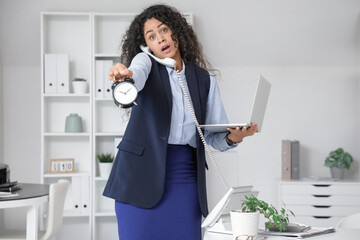 Wall Mural - Young African-American businesswoman with alarm clock and laptop trying to meet deadline in office