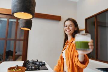 Canvas Print - Young woman smiling while holding a green smoothie in a modern kitchen, wearing an orange jacket and conveying a healthy lifestyle