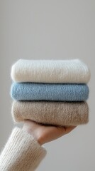 Poster - A hand cradles a stack of three soft knitwear pieces in calming shades of light blue and beige, inviting warmth and comfort during a peaceful autumn afternoon