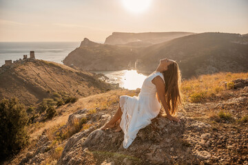 Wall Mural - A woman in a white dress is sitting on a rock overlooking a body of water. She is enjoying the view and taking in the scenery.