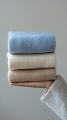 Poster - A hand gently cradles a stack of three cozy sweaters in light blue, beige, and cream, bringing warmth and comfort to any chilly day while displaying a minimalist aesthetic