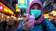 © DKPhoto - A young woman wearing a face mask holds a bottle of hand sanitizer in a bustling city street, representing safety and health in an urban environment during pandemic times.
