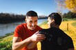 © Studio Marmellata - Two men in casual activewear laugh and interact cheerfully near a scenic river trail surrounded by vibrant autumn foliage under the bright sun
