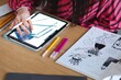 © Studio Marmellata - A close-up of a person using a stylus to draw a colorful design on a tablet screen, with colored pens and a comic book on a wooden desk in a creative workspace