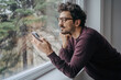 © Akbari - Young handsome middle eastern man scrolling on mobile phone in the morning standing near window.