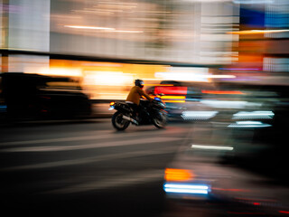 Motorcycle with rider in helmet riding through bright Tokyo city lights at night quickly. Speeding or in a rush amongst traffic and urban buildings with vibrant colours and panning motion blur