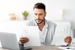 © Prostock-studio - Millennial male self-entrepreneur working with papers at desk at home office, checking annual financial reports, free space. Young man reading documents, sitting at workplace with computer