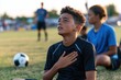 © Milos - A young boy holds his chest in distress while sitting on the soccer field, reflecting feelings of discomfort amid a lively practice session, highlighting sports and youth.