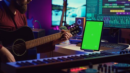 Leinwandbilder - Male artist learning to play guitar on online lesson tutorial, practicing acoustic instrument and looking at phone with greenscreen display. Music producer doing rehearsal guitar play. Camera A.