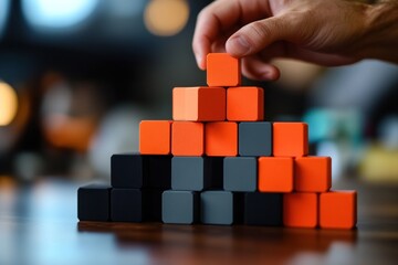Poster - Hand placing orange block atop pyramid of orange and grey blocks on wooden table.