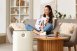 © Pixel-Shot - Modern air purifier against woman with magazine sitting on sofa at home, closeup