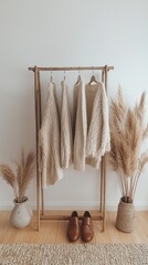 Leinwandbilder - In a cozy corner, a rack displays soft neutral-colored sweaters, harmonizing with minimalistic decor. Drying pampas grass stands elegantly beside, inviting warmth and comfort