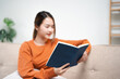 © Fahng - girl enjoying an interesting novel reading in living room . Hobbies and activities. Happy young asian woman reading book, sitting on sofa lady enjoying interesting story, studying for exam,