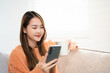 © Fahng - Asian woman with smile use tablet smartphone in orange winter sweater work home, beauty asian girl hygge relax in living room. Technology people connection digital online social media market banner