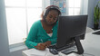 © Krakenimages.com - Young african american woman working in an office, writing notes in front of a computer, wearing headphones with a green sweater in a modern indoor workspace