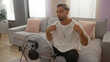 © Krakenimages.com - Young man with a beard seated in an apartment's living room, cooling off in front of a fan, wearing a white shirt, with pillows on the couch and natural light coming in through the curtains.