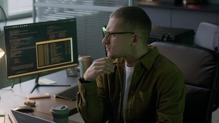 Medium close up of young concentrated Caucasian male programmer in eyeglasses and casual clothes sipping coffee while working in evening office with program coding on pc computer in blurred background