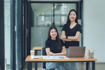 Confident Co-workers: Portrait of two smiling Asian female entrepreneurs sitting at a modern office desk with a laptop, showcasing teamwork and success. 