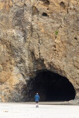 Wall Mural - Man walks along a sandy beach towards a large cave in a cliff face. Coastal scenery. BETHELLS BEACH, WAITAKERES, AUCKLAND, NEW ZEALAND