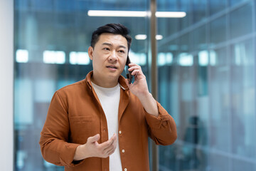 Poster - Asian man mature businessman in an office setting, engaged in a conversation using a smartphone. The image captures a professional moment, reflecting communication, business, and technology use