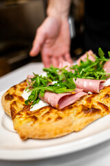 Wall Mural - freshly baked pizza topped with slices of ham and arugula, served on a white plate. A chef's hand is positioned in the background, enhancing the culinary presentation. Ideal for food-related content.