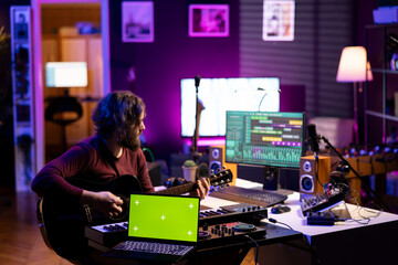 Leinwandbilder - Sound engineer playing guitar next to laptop showing greenscreen display, recording songs with mixing console and daw software interface. Producer composes new music on acoustic instrument.
