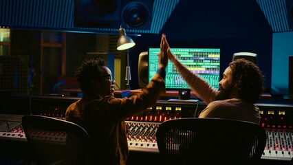 Fototapete - Musician and audio engineer sharing high five after creating a beautiful hit song, enjoying success in professional studio. Team of producer an artist celebrating teamwork in control room. Camera B.