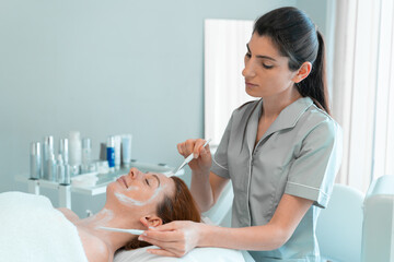 Wall Mural - Professional masseuse applying face mask with brush on woman's face at cosmetology salon. Relaxed female customer is getting pampered in spa by beautician. 