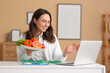 © Pixel-Shot - Young female nutritionist with healthy food video chatting at table in office